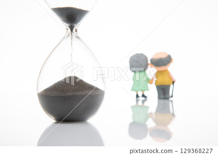 two elderly figurines holding hands, gazing at a large hourglass with most sand in the top 129368227