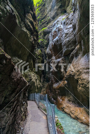 Narrow walkway carved into the cliffside along a turquoise stream inside Partnachklamm Gorge, Garmisch-Partenkirchen, Bavaria. A stunning natural passageway through dramatic alpine rock formations. 129369383