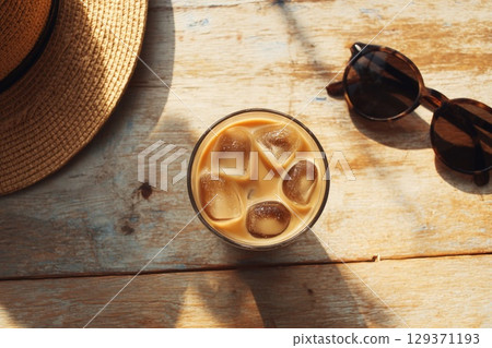 cold coffee drink with ice cubes in an iced latte glass, placed on the wooden table next to sunglasses and a sun hat. cold coffee drink with ice cubes in an iced latte glass, placed on the wooden table next to sunglasses and a sun hat. 129371193