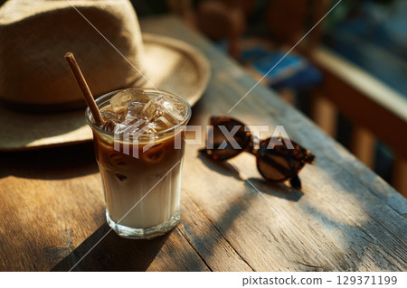 cold coffee drink with ice cubes in an iced latte glass, placed on the wooden table next to sunglasses and a sun hat. cold coffee drink with ice cubes in an iced latte glass, placed on the wooden table next to sunglasses and a sun hat. 129371199