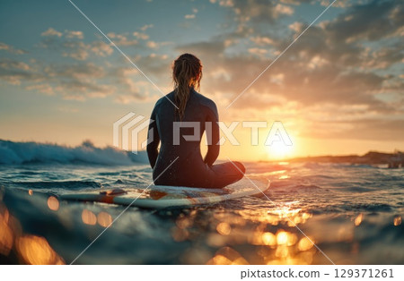 A female surfer in her wetsuit, sitting on a surfboard at sea with a sunset in the background 129371261