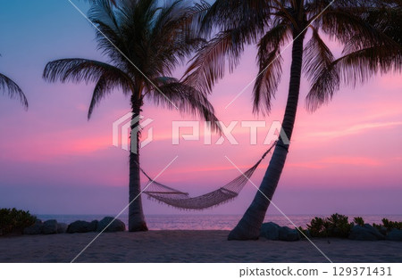 A cozy hammock between two palm trees on the beach at sunset, with a pink and purple sky. 129371431