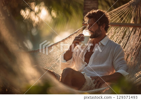handsome man with a beard sitting in a hammock, drinking milk in a summer tropical environment 129371493