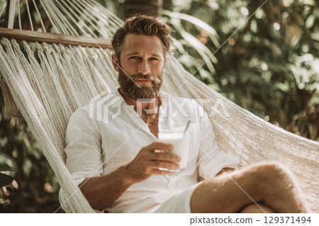 handsome man with a beard sitting in a hammock, drinking milk in a summer tropical environment handsome man with a beard sitting in a hammock, drinking milk in a summer tropical environment 129371494