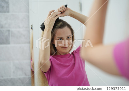 A woman is expertly styling her hair using a blow dryer and brush while in the bathroom A woman is expertly styling her hair using a blow dryer and brush while in the bathroom 129373911