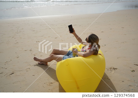 A young woman on the beach lounging on a yellow bean bag  129374568