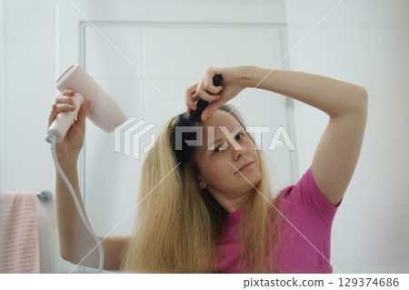 A woman is expertly styling her hair using a blow dryer and brush while in the bathroom A woman is expertly styling her hair using a blow dryer and brush while in the bathroom 129374686
