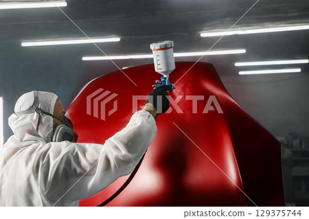 Professional spray painter in protective mask and clothes spraying car hood of vehicle part to red color using spray gun in paint chamber. Professional spray painter in protective mask and clothes spraying car hood of vehicle part to red color using spray gun in paint chamber. 129375744