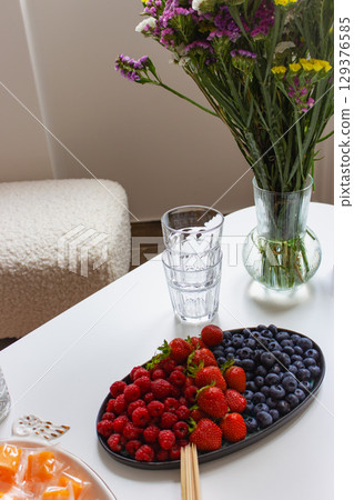 Blueberries, strawberries and raspberries on plate with wooden sticks on white table. Summer berries and flowers in vase. Summer harvest. Sweet berries and fruits. Healthy food. Natural lifestyle. Blueberries, strawberries and raspberries on plate with wooden sticks on white table. Summer berries and flowers in vase. Summer harvest. Sweet berries and fruits. Healthy food. Natural lifestyle. 129376585