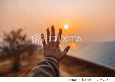 Hand Reaching Out to Touch Glowing Solar Panel Under Beautiful Sunset Sky 129377380