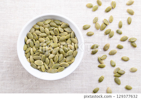Pepitas, hulled pumpkin seeds, in a white bowl, on linen. Raw, dried squash seeds, fruits of Cucurbita, light green after the husk is removed, and used as snack, garnish or as an ingredient. Photo Pepitas, hulled pumpkin seeds, in a white bowl, on linen. Raw, dried squash seeds, fruits of Cucurbita, light green after the husk is removed, and used as snack, garnish or as an ingredient. Photo 129377632