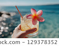 hand holding a tropical cocktail with a straw and a pink plumeria flower on a beach 129378352