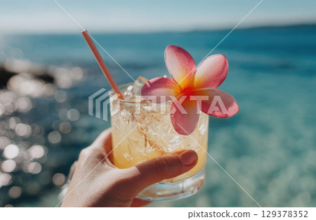 hand holding a tropical cocktail with a straw and a pink plumeria flower on a beach hand holding a tropical cocktail with a straw and a pink plumeria flower on a beach 129378352