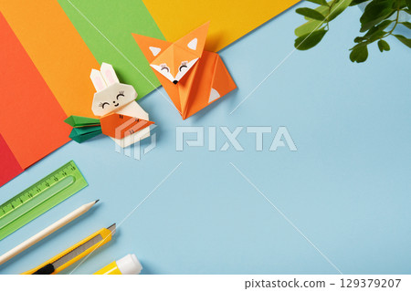 Origami fox and rabbit with carrot with colorful paper and craft tools on blue background. Copyspace. Concept of handmade art and creativity. 129379207