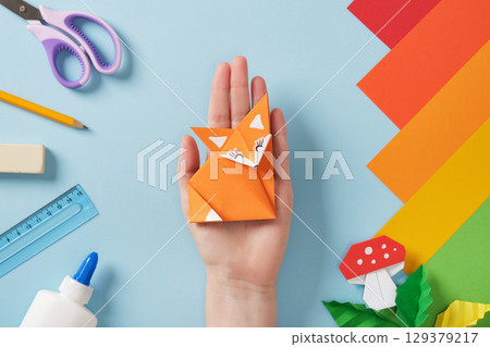 Hand holding origami fox. Colorful paper and craft tools on a blue background. Concept of handmade art and creativity. Hand holding origami fox. Colorful paper and craft tools on a blue background. Concept of handmade art and creativity. 129379217