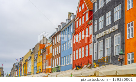 Vibrant, historic buildings line the canals of Nyhavn in Copenhagen, showcasing a mix of colors and architectural styles. Boats rest nearby, creating a lively atmosphere for visitors. 129380001