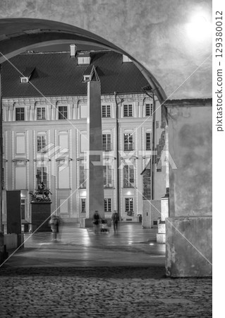 The illuminated archway of a covered bridge leads to Saint Vitus Cathedral in Prague, casting shadows on cobblestones as visitors stroll through. The atmosphere is tranquil and historic. 129380012