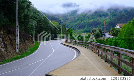 A winding asphalt road curves through a lush, green mountainous landscape on a cloudy day A winding asphalt road curves through a lush, green mountainous landscape on a cloudy day 129383555