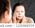 Female applying moisturizing face mask in bathroom. Beauty routine. International Self Care Day. 129384559