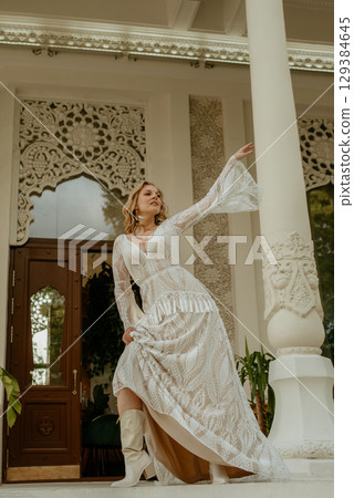 Beautiful slim blonde poses in boho wedding dresses and an embroidered corset on a summer terrace 129384645
