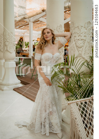 Beautiful slim blonde poses in boho wedding dresses and an embroidered corset on a summer terrace Beautiful slim blonde poses in boho wedding dresses and an embroidered corset on a summer terrace 129384663