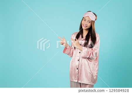 Happy young woman in pajama sleep mask pointing finger, eye open relaxing at home, calm and relaxed she is pointing to a copy space area for wellness designs, headshots isolated studio blue background 129384928