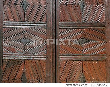 Close-up of a rustic wooden door featuring symmetrical geometric patterns and nail detailing. Traditional craftsmanship, architectural texture, woodworking design, cultural heritage 129385847