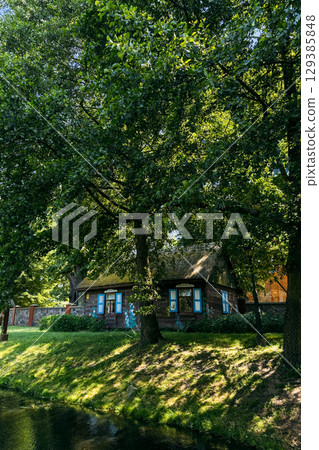 Traditional thatched-roof wooden cottage with blue window shutters shaded by large trees near a calm pond. Cultural preservation, rural housing, vernacular architecture, ecological living Traditional thatched-roof wooden cottage with blue window shutters shaded by large trees near a calm pond. Cultural preservation, rural housing, vernacular architecture, ecological living 129385848