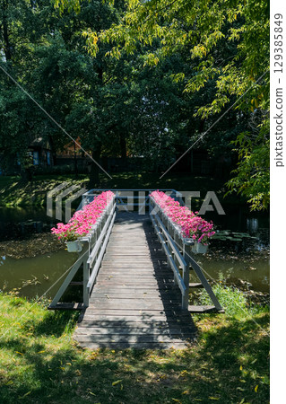 Wooden footbridge decorated with rows of pink flowers crossing a calm pond in a shaded garden. Landscape design, floral decoration, outdoor leisure, serene environment Wooden footbridge decorated with rows of pink flowers crossing a calm pond in a shaded garden. Landscape design, floral decoration, outdoor leisure, serene environment 129385849