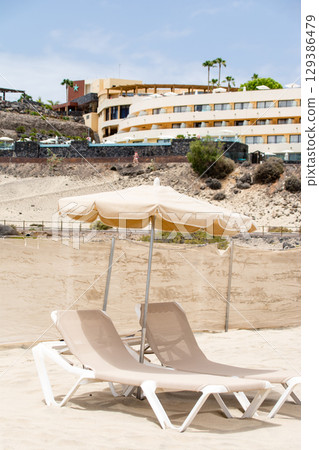 Beach beds lined up on a spacious sandy beach and a coastal resort hotel on Fuerteventura Island 129386479
