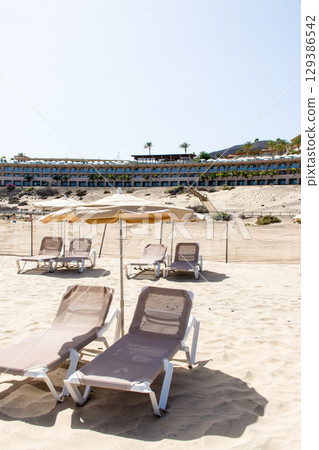 Beach beds lined up on a spacious sandy beach and a coastal resort hotel on Fuerteventura Island 129386542