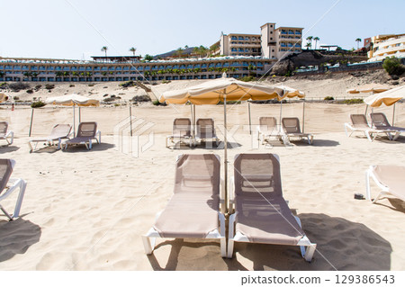 Beach beds lined up on a spacious sandy beach and a coastal resort hotel on Fuerteventura Island 129386543