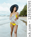 woman is walking on the beach wearing  yellow bikini and  black hat 129387095