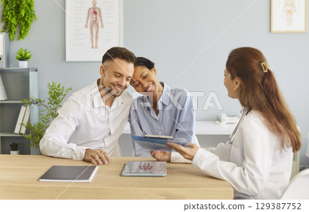 Smiling happy young couple sitting with female family doctor therapist in medical clinic. 129387752