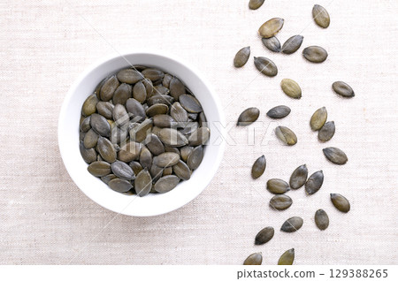 Styrian oil pumpkin seeds, in a white bowl, on linen. Dried, dark green and young fruits of the Styrian pumpkin, Cucurbita pepo var. styriaca, a variety of the common pumpkin, cultivated for seed oil. 129388265
