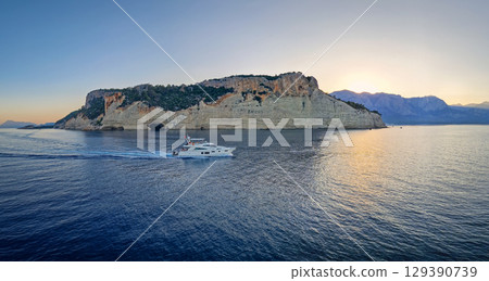 Evening seascape panorama with a boat cruising away on the blue sea, near the Pirate Cave in Kemer at sunset. Beautiful Turkish mediterranean coastline lined with tree covered rocky cliffs at dusk Evening seascape panorama with a boat cruising away on the blue sea, near the Pirate Cave in Kemer at sunset. Beautiful Turkish mediterranean coastline lined with tree covered rocky cliffs at dusk 129390739