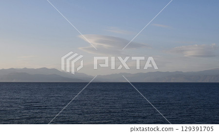 Beautiful coastal view showcasing vast ocean expanse meeting distant mountain range during bright daylight, soft clouds drifting above creating serene atmosphere 129391705