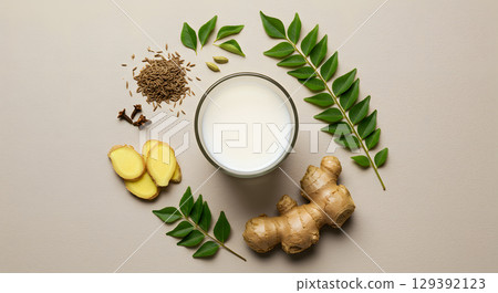 Fresh ginger root and slices with green leaves and spices surrounding glass of milk 129392123