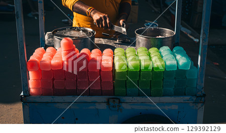 Colorful ice cubes stacked on street vendor cart with person preparing food behind 129392129