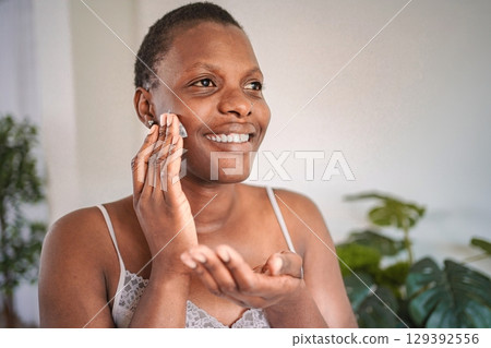 Young woman applying moisturizer on her face smiling at camera Young woman applying moisturizer on her face smiling at camera 129392556