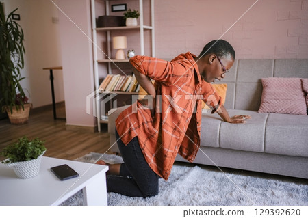 Young woman suffering back pain at home, kneeling on the floor Young woman suffering back pain at home, kneeling on the floor 129392620