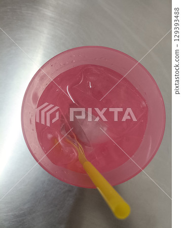 Pink plastic cup filled with ice cubes and yellow straw, placed on metallic surface, creating refreshing and colorful beverage presentation 129393488