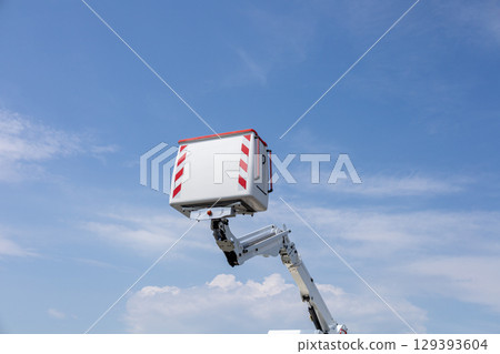 A hydraulic lift is raised high, showcasing its platform against a bright blue sky with scattered clouds, indicating outdoor work activity 129393604