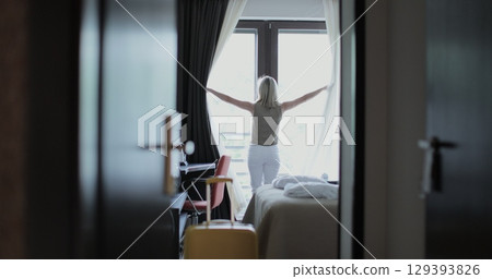 Young blond woman traveler entering sunlit hotel room, walking toward window with suitcase and unveil curtains from window. Embodying travel anticipation. 129393826