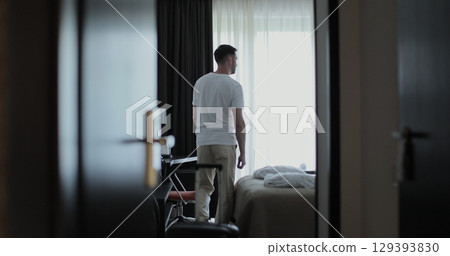Male traveler entering sunlit hotel room, walking toward window with suitcase and unveil curtains from window. Embodying travel anticipation. 129393830