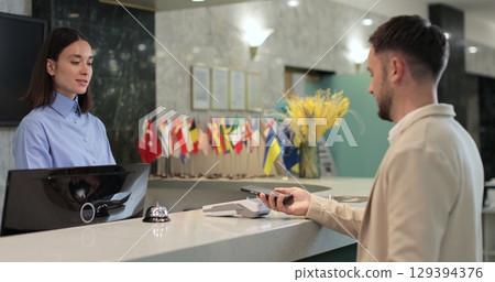 Man makes nfc payment with mobile phone at pos terminal, paying for room at hotel. Person at reception desk and doing check. Man makes nfc payment with mobile phone at pos terminal, paying for room at hotel. Person at reception desk and doing check. 129394376