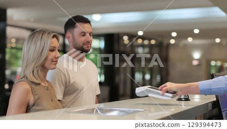 Woman makes nfc payment with mobile phone at pos terminal, paying for room at hotel. Person at reception desk and doing check. 129394473