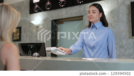 Woman makes nfc payment with mobile phone at pos terminal, paying for room at hotel. Person at reception desk and doing check. 129394502