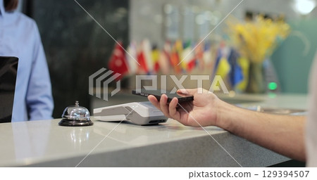 Customer with mobile phone pay for hotel reception, payment. Client, concierge service or people at help desk. Person at reception desk and doing check. Close up. Customer with mobile phone pay for hotel reception, payment. Client, concierge service or people at help desk. Person at reception desk and doing check. Close up. 129394507
