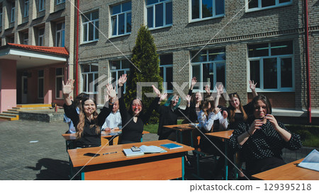 Happy students sitting at desks outside school building, raising hands and having a great time on a school day Happy students sitting at desks outside school building, raising hands and having a great time on a school day 129395218